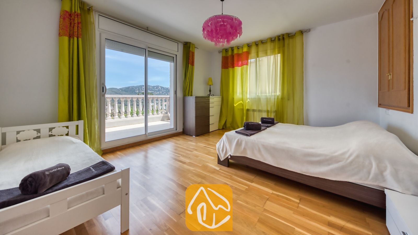 Vakantievilla's Costa Brava Spanje - Villa Primavera - Slaapkamer met twee bedden, houten vloer, groene gordijnen en een glazen schuifpui die uitkomt op een balkon met uitzicht op de heuvels.