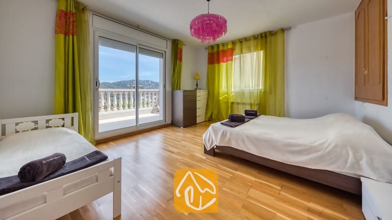 Villas de vacances Costa Brava Espagne - Villa Primavera - Chambre avec deux lits, parquet en bois, rideaux verts et porte-fenêtre coulissante s'ouvrant sur un balcon offrant une vue sur les collines.