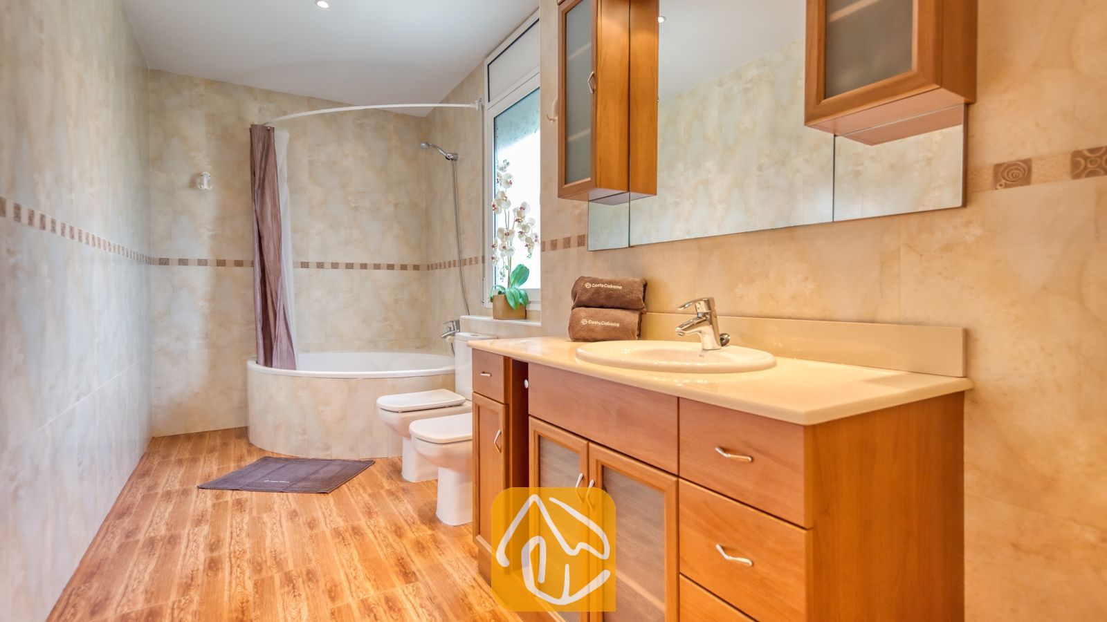 Vakantievilla's Costa Brava Spanje - Villa Primavera - Badkamer voorzien van een houten badkamermeubel met lattes, een aangeronde ligbad met een douchegordijn en beige betegelde muren.