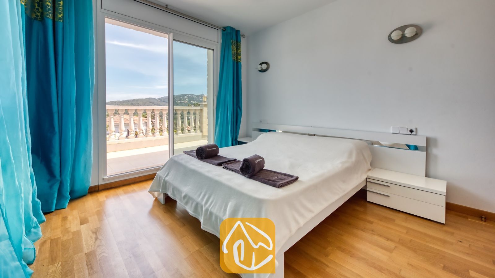 Vakantievilla's Costa Brava Spanje - Villa Primavera - Slaapkamer met een wit bed, houten vloeren, helderblauwe gordijnen en glazen schuifdeuren die opengaan naar een balkon met balustrades.