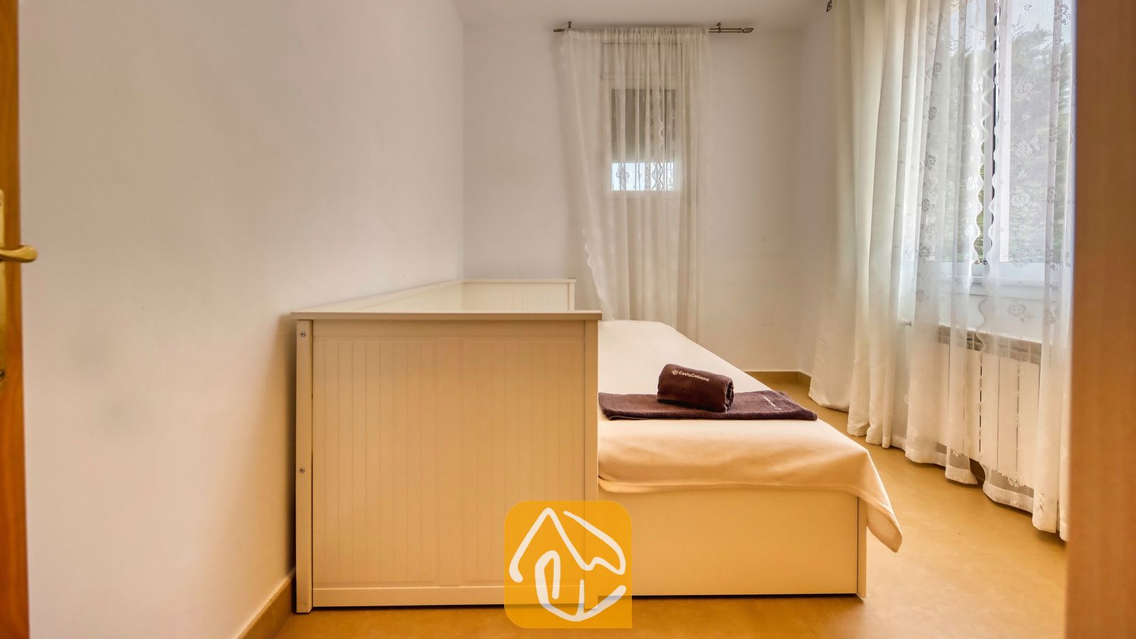 Vakantievilla's Costa Brava Spanje - Villa Primavera - Slaapkamer met een wit houten eenpersoonsdivanbed, opgevouwen bruine handdoeken en doorschijnende witte gordijnen voor twee ramen.