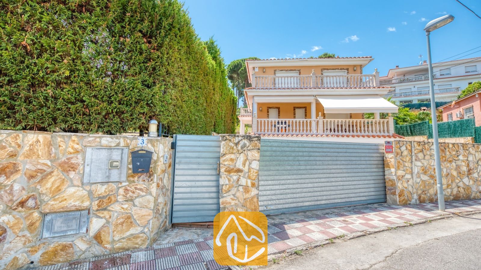 Vakantievilla's Costa Brava Spanje - Villa Primavera - Buitenaanzicht van Villa Primavera met een stenen erfafscheiding, metalen toegangspoorten en een beige huis van twee verdiepingen met witte balkons voorzien van balustrades.