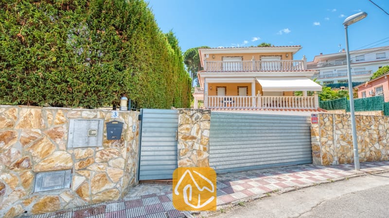 Villas de vacances Costa Brava Espagne - Villa Primavera - Vue extérieure de la Villa Primavera montrant un muret en pierre, un portail d'entrée en métal et une maison beige à deux étages dotée de balcons avec balustrades blanches.