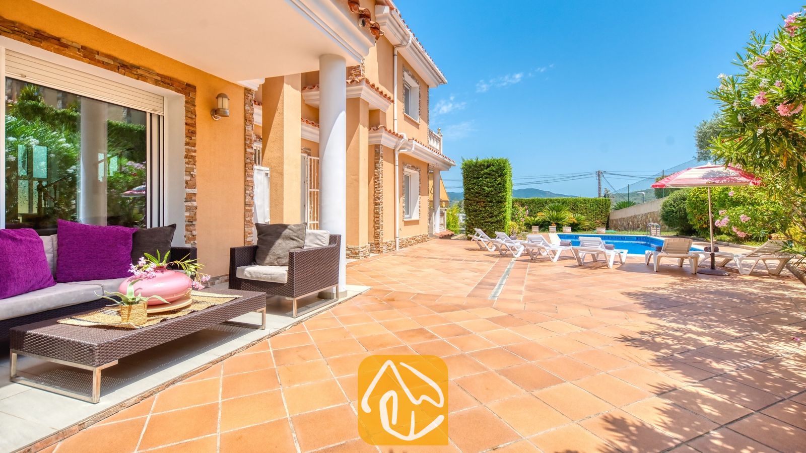 Vakantievilla's Costa Brava Spanje - Villa Primavera - Overdekt terras met rieten bank en roze kussens bij Villa Primavera, met uitzicht op een betegeld terras en een buitenzwembad onder een blauwe lucht.