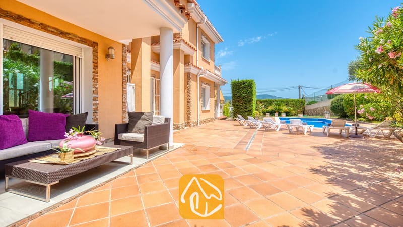 Vakantievilla's Costa Brava Spanje - Villa Primavera - Overdekt terras met rieten bank en roze kussens bij Villa Primavera, met uitzicht op een betegeld terras en een buitenzwembad onder een blauwe lucht.