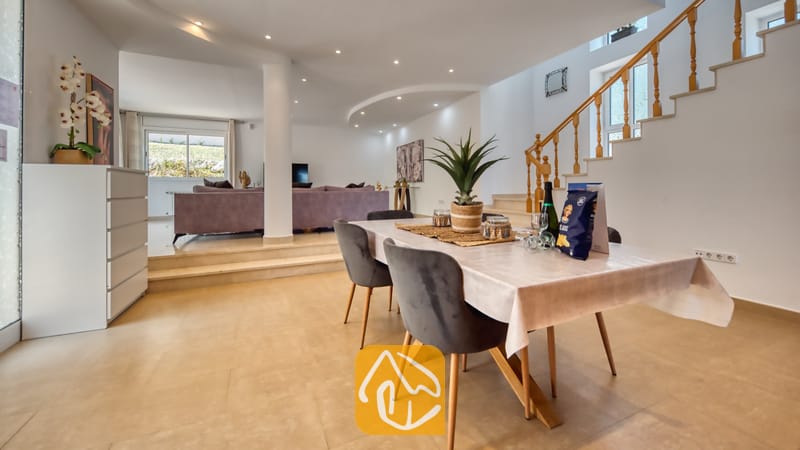 Vakantievilla's Costa Brava Spanje - Villa Primavera - Eethoek met een houten tafel en grijsvelours stoelen bij Villa Primavera, met op de achtergrond een trap met houten leuningen en een verzonken woonkamer.