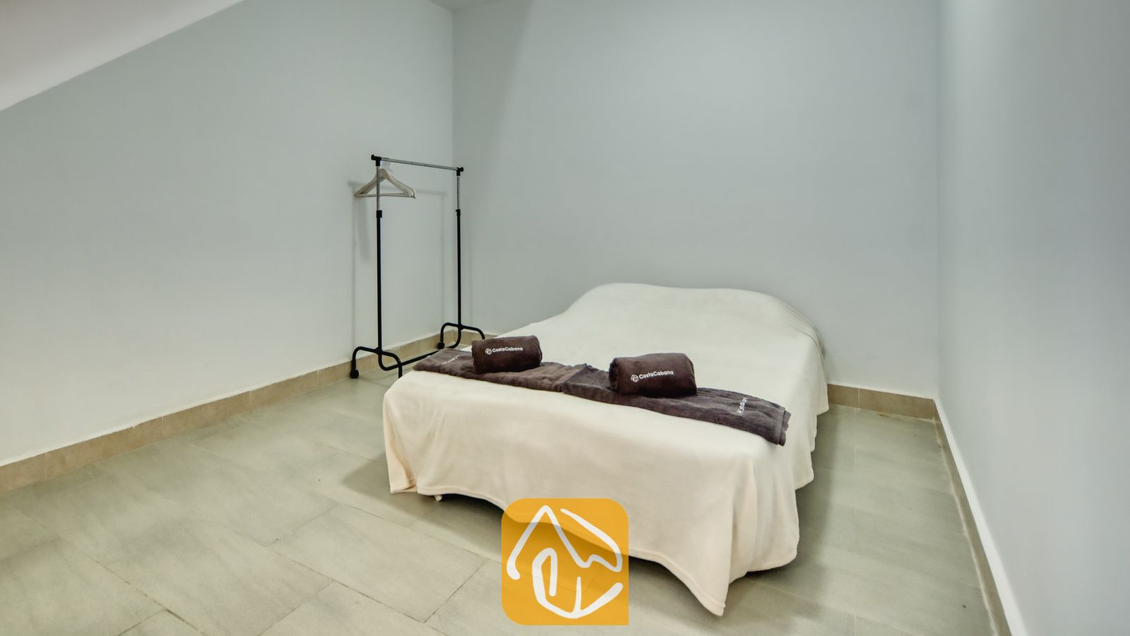 Vakantievilla's Costa Brava Spanje - Villa Primavera - Slaapkamer met een tweepersoonsbed opgemaakt in crèmekleurige lakens, opgerolde donkerbruine handdoeken en een metalen kledingrek tegen een lichtgrijze muur.