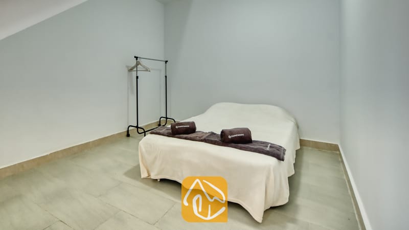 Villas de vacances Costa Brava Espagne - Villa Primavera - Chambre avec un lit double habillé de linge de lit crème, des serviettes marron foncé enroulées et un portant à vêtements en métal adossé à un mur gris pâle.