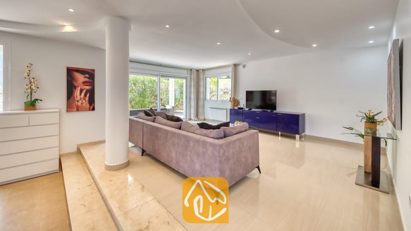 Vakantievilla's Costa Brava Spanje - Villa Primavera - Ruime woonkamer in Villa Primavera met een paarse hoekbank, een hoogglans blauwe tv-meubel en grote glazen schuifdeuren die opengaan naar een tuin.