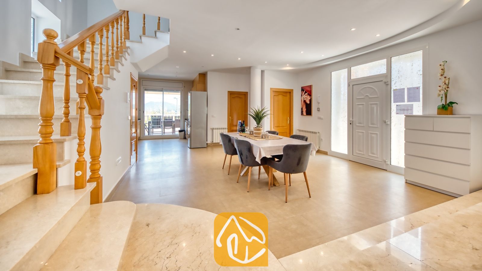 Vakantievilla's Costa Brava Spanje - Villa Primavera - Open interieur van Villa Primavera met een marmeren trap met houten leuning, een eettafel met grijs gestoffeerde stoelen en witte tegelvloeren.
