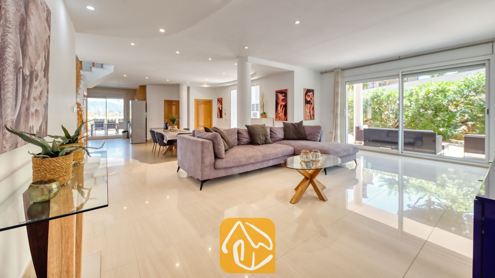 Vakantievilla's Costa Brava Spanje - Villa Primavera - Ruime woonkamer in Villa Primavera met een grote grijze hoekbank, glanzende tegelvloeren, een glazen salontafel en grote glazen schuifdeuren die uitkomen op een tuin.