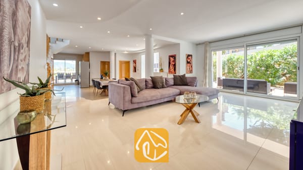 Vakantievilla's Costa Brava Spanje - Villa Primavera - Ruime woonkamer in Villa Primavera met een grote grijze hoekbank, glanzende tegelvloeren, een glazen salontafel en grote glazen schuifdeuren die uitkomen op een tuin.