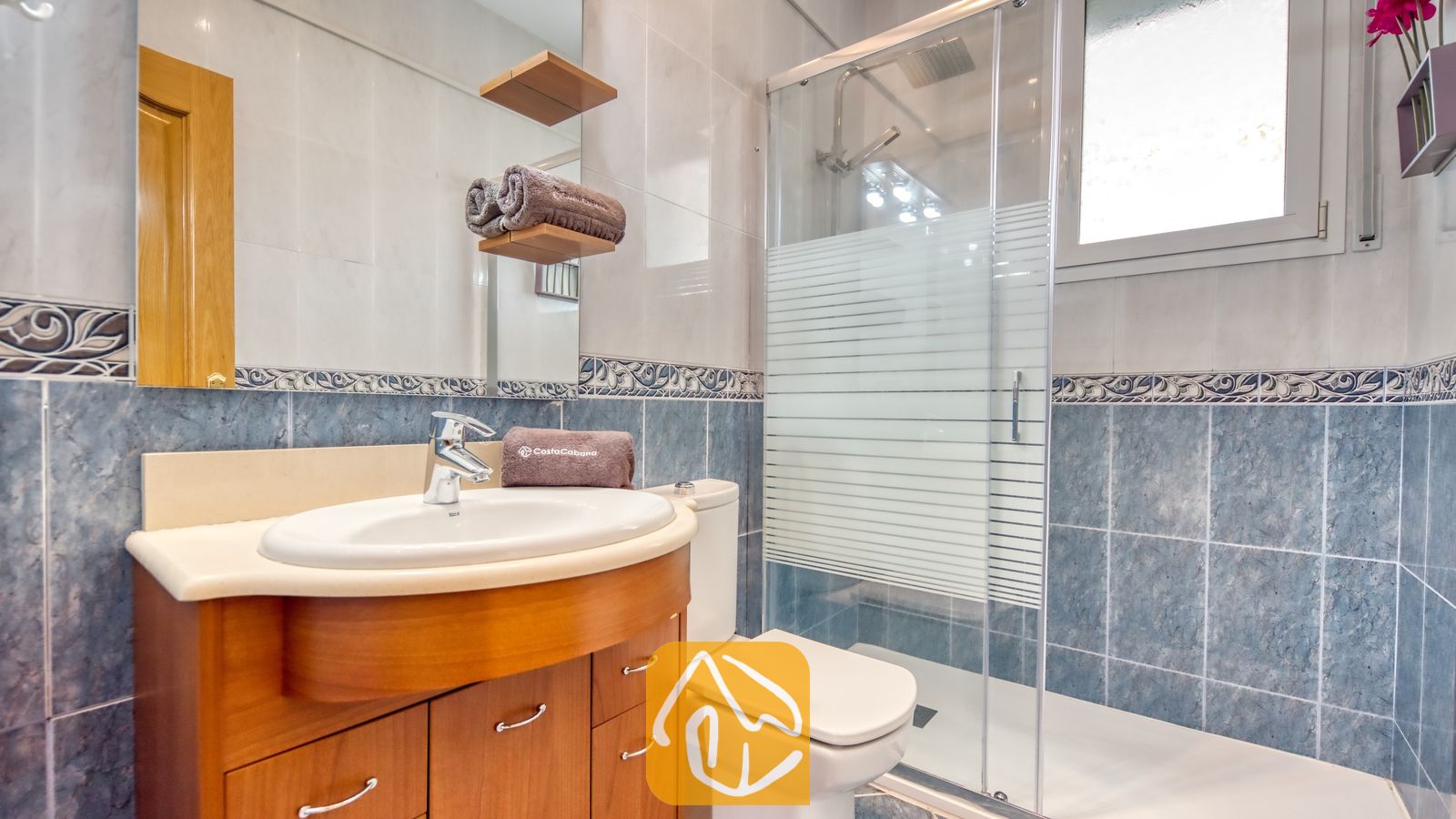 Vakantievilla's Costa Brava Spanje - Villa Primavera - Moderne badkamer met blauwe tegels op de wanden, een houten badkamermeubel met een witte wastafel, een toilet en een glazen inloopdouche.
