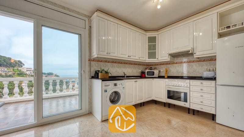 Vakantievilla's Costa Brava Spanje - Villa Sunrise - Keuken voorzien van witte kasten, een ingebouwde oven, een wasmachine en een glazen schuifdeur die toegang biedt tot een buitenbalkon met uitzicht op de verre heuvels en de zee.