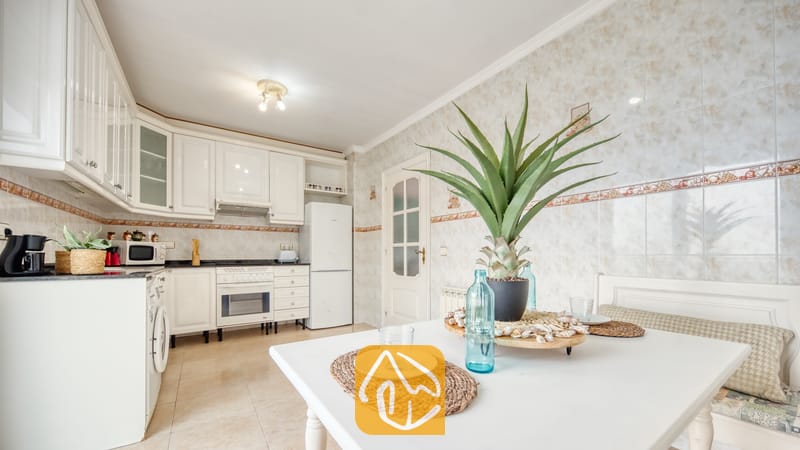 Vakantievilla's Costa Brava Spanje - Villa Sunrise - Witte eettafel met geweven placemats, glaswerk en een potplant in een keuken met witte kasten en tegelmuren met een patroon.