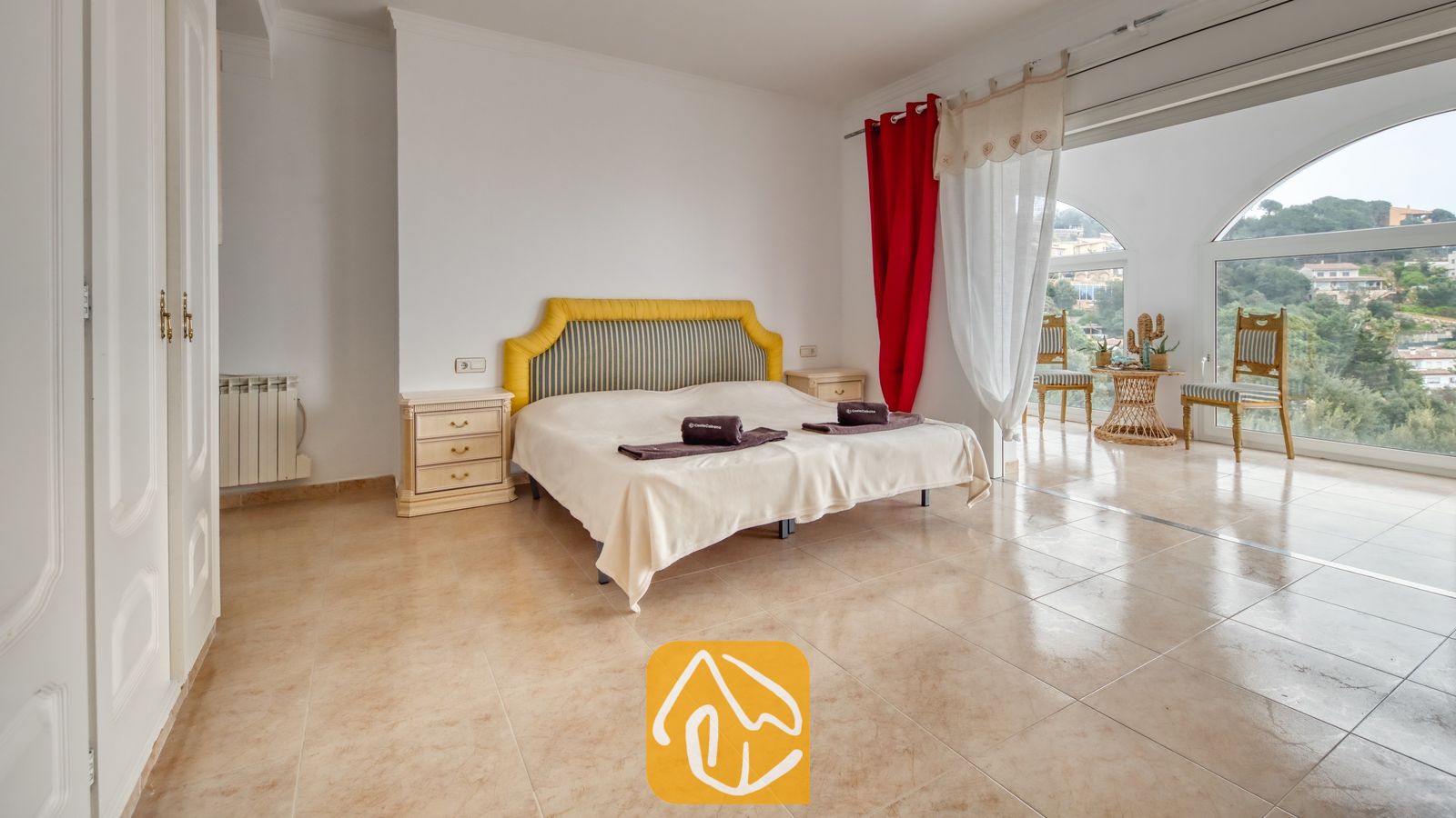 Vakantievilla's Costa Brava Spanje - Villa Sunrise - Slaapkamer met een tweepersoonsbed voorzien van een geel gestreept hoofdbord, tegelvloeren en grote boogvormige ramen met uitzicht op de heuvels.