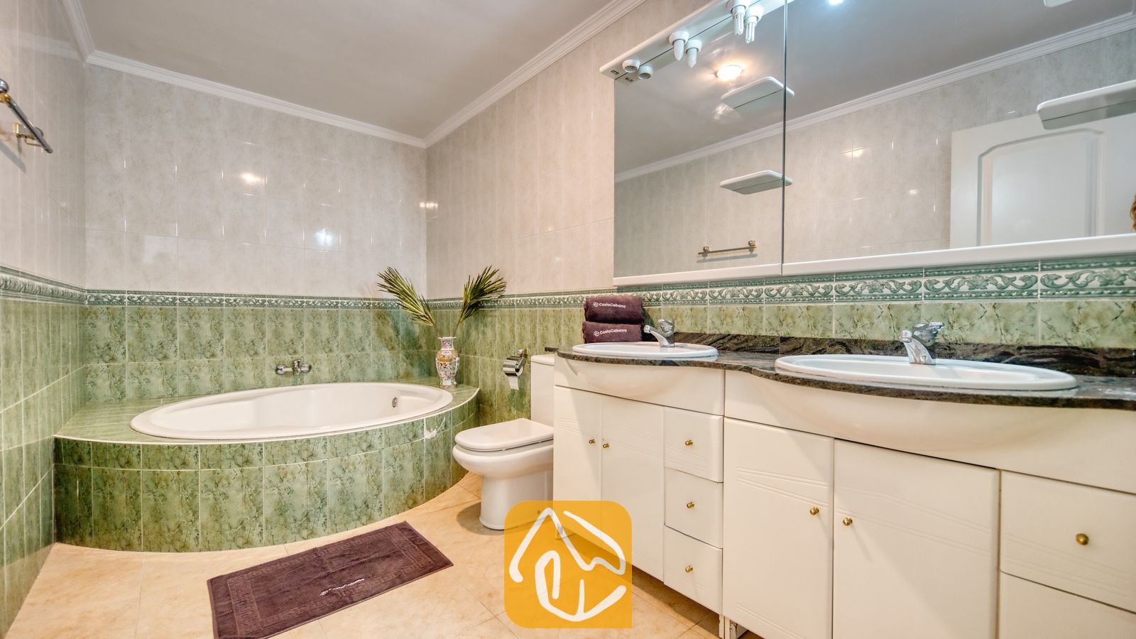 Vakantievilla's Costa Brava Spanje - Villa Sunrise - Badkamer met een groen getegeld ovaal ligbad, een wit dubbel meubel met twee wastafels en een grote wandspiegel.