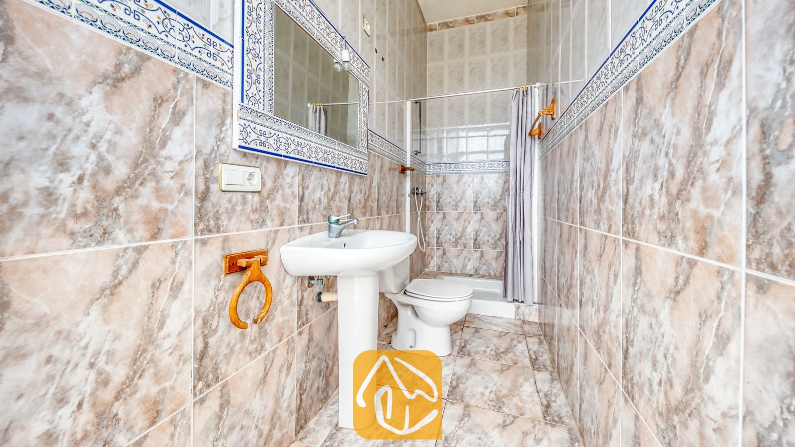 Vakantievilla's Costa Brava Spanje - Villa Sunrise - Badkamer voorzien van bruine marmeren tegels, een witte wastafel op zuil met een bijpassende spiegel, en een toilet naast een inloopdouche met een grijs gordijn.