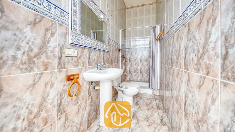 Holiday villas Costa Brava Spain - Villa Sunrise - Bathroom featuring marbled brown tiles, a white pedestal sink with a matching mirror, and a toilet next to a walk-in shower with a grey curtain.