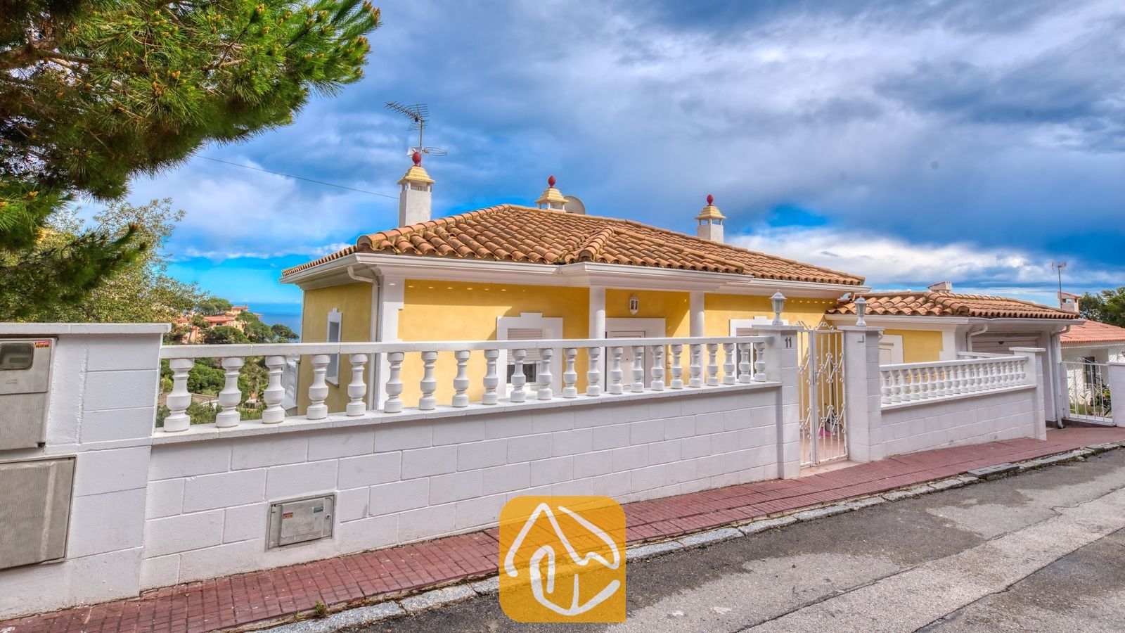 Vakantievilla's Costa Brava Spanje - Villa Sunrise - Buitenaanzicht van Villa Sunrise met de gele gevel, het dak van terracotta pannen en een witte balustrade langs de straat.