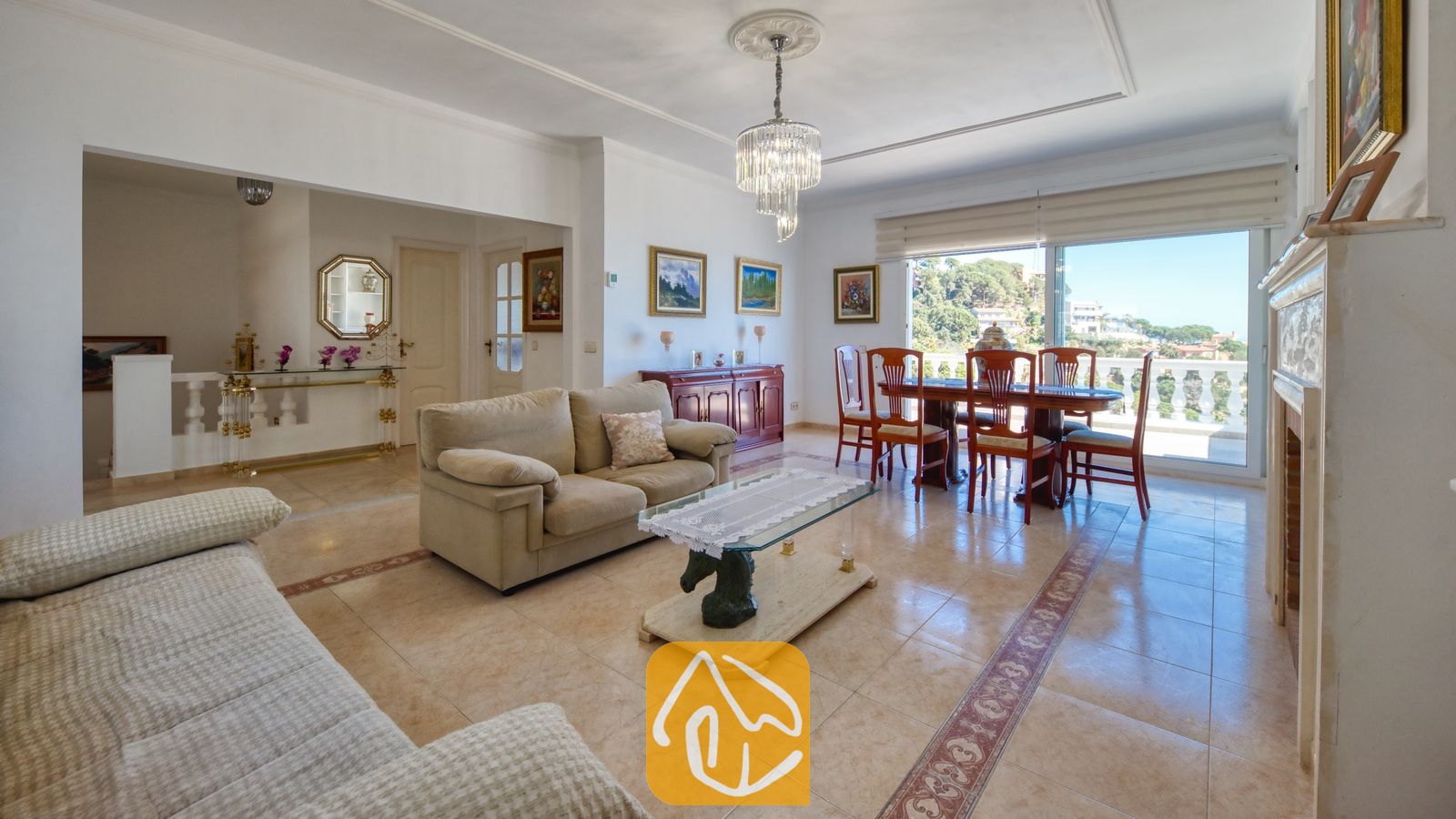 Vakantievilla's Costa Brava Spanje - Villa Sunrise - Ruime woon- en eetkamer met plavuizen, beige banken, een glazen salontafel met een paardensculptuur als voet en een kristallen kroonluchter in Villa Sunrise.