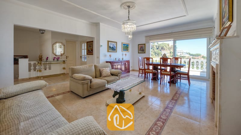 Vakantievilla's Costa Brava Spanje - Villa Sunrise - Ruime woon- en eetkamer met plavuizen, beige banken, een glazen salontafel met een paardensculptuur als voet en een kristallen kroonluchter in Villa Sunrise.