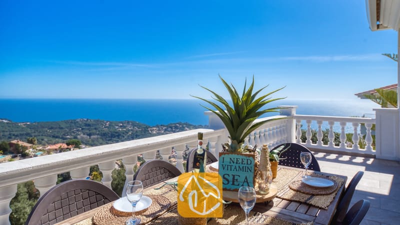 Vakantievilla's Costa Brava Spanje - Villa Sunrise - Terras met uitzicht op zee bij Villa Sunrise, voorzien van een houten eettafel, witte borden en een decoratief bord te midden van beplante potten.