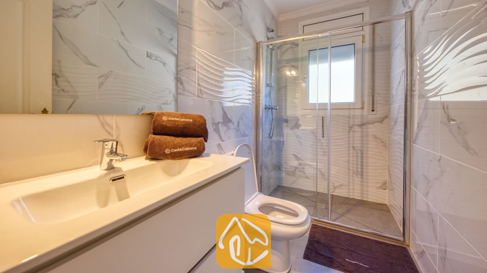 Vakantievilla's Costa Brava Spanje - Villa Sunrise - Moderne badkamer met wit marmeren tegelwanden, een wit wastafelmeubel, een toilet en een glazen inloopdouche, met bruine handdoeken opgestapeld op het aanrecht.
