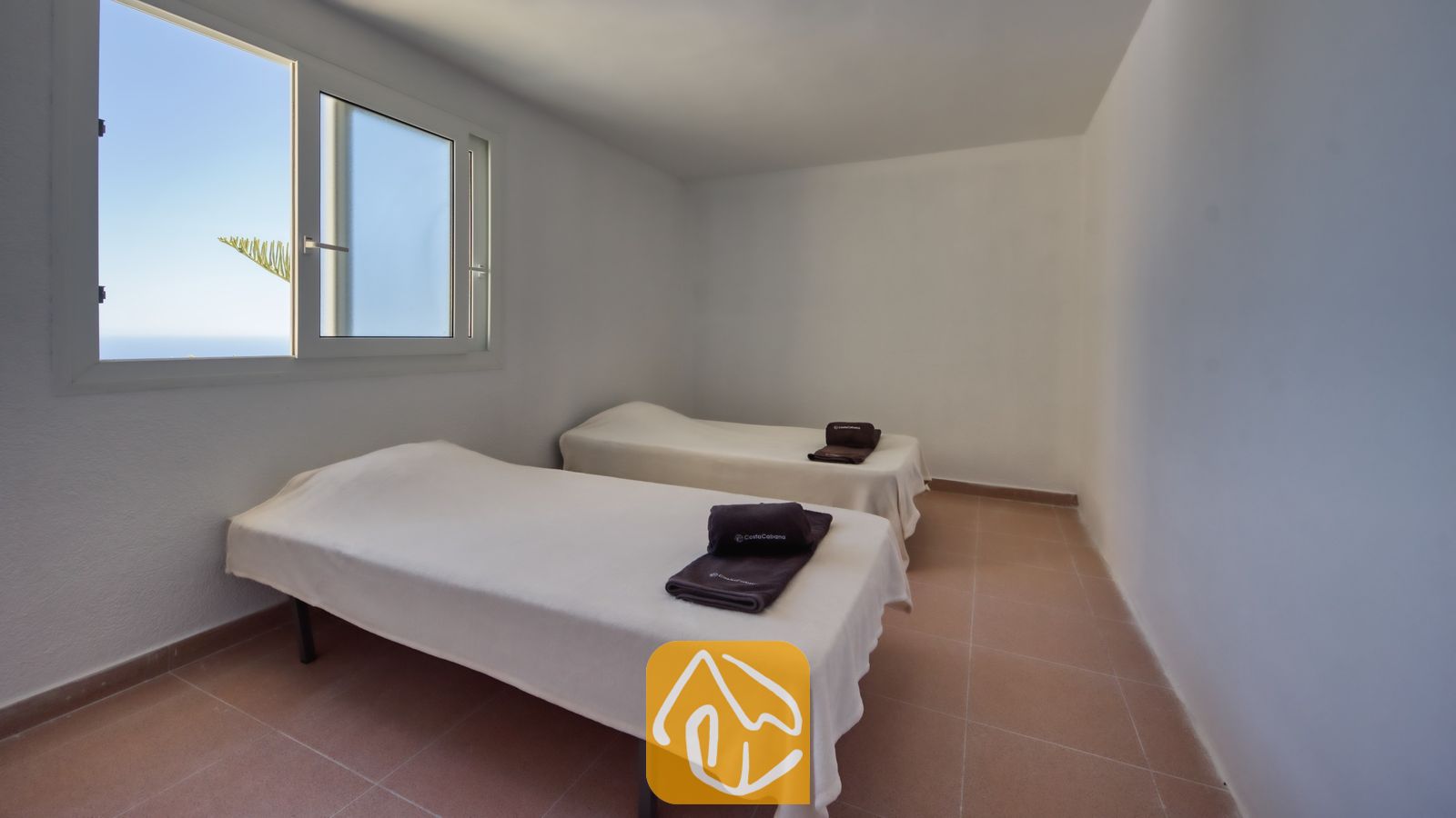 Vakantievilla's Costa Brava Spanje - Villa Sunrise - Slaapkamer met twee eenpersoonsbedden opgemaakt met witte overtrekken, donkere opgevouwen handdoeken, tegelvloeren en een raam met uitzicht op een blauwe hemel.