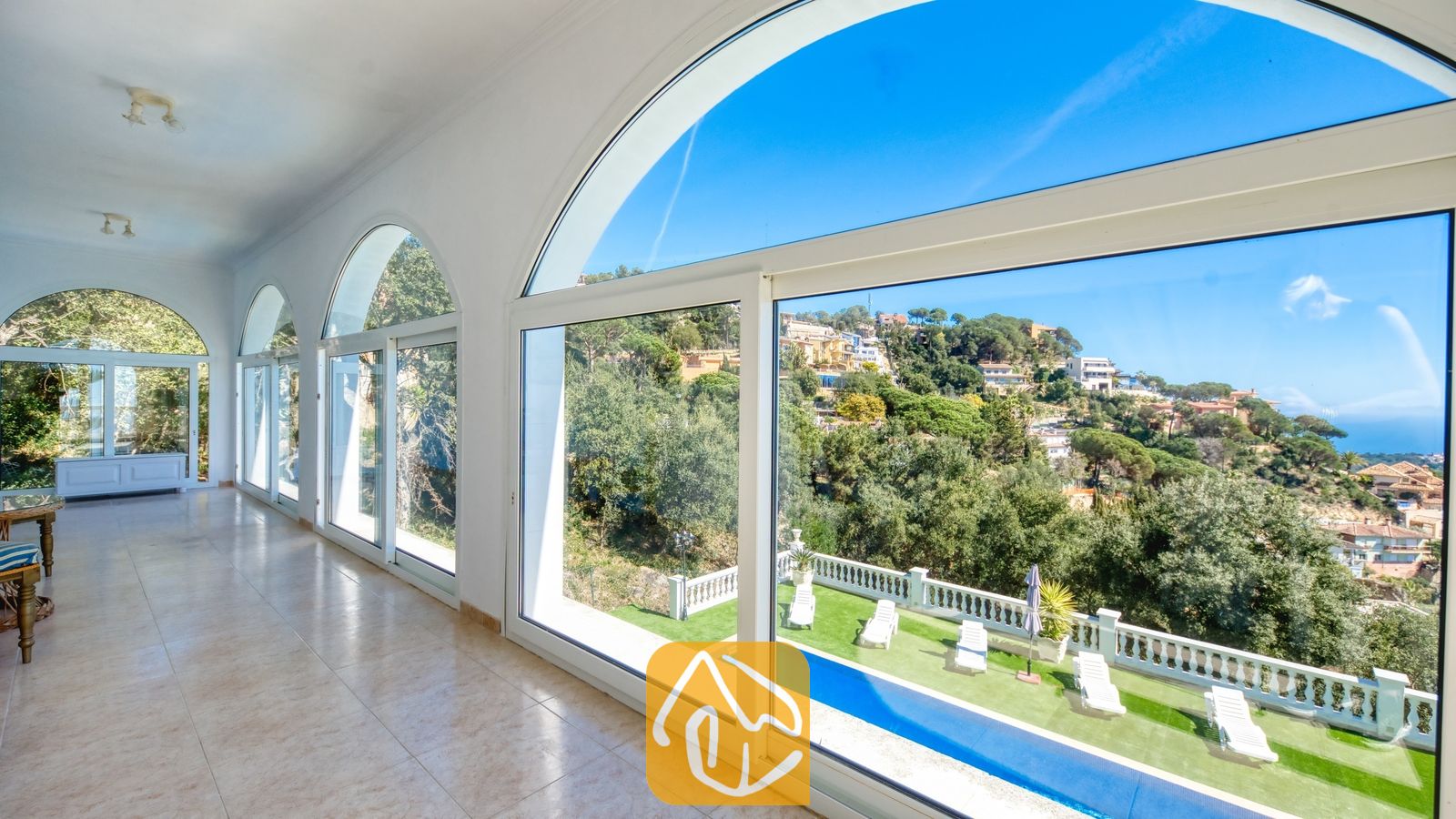 Vakantievilla's Costa Brava Spanje - Villa Sunrise - Ruime binnenhal van Villa Sunrise, voorzien van grote boogvormige ramen met uitzicht op een heuvelachtig landschap en een zwembad.