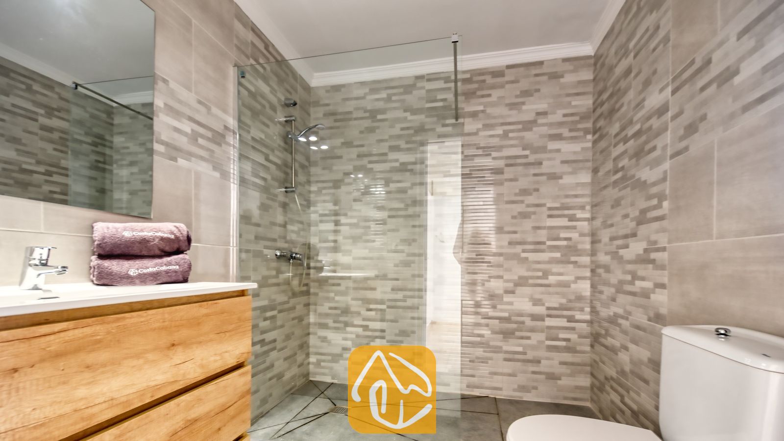 Vakantievilla's Costa Brava Spanje - Villa Sunrise - Moderne badkamer met grijze mozaïektegels op de muren, een glazen inloopdouche, een houten badkamermeubel met een witte wastafel en paarse handdoeken.
