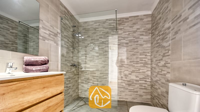 Holiday villas Costa Brava Spain - Villa Sunrise - Modern bathroom featuring grey mosaic tiled walls, a walk-in glass shower enclosure, a wooden vanity with a white sink, and purple towels.