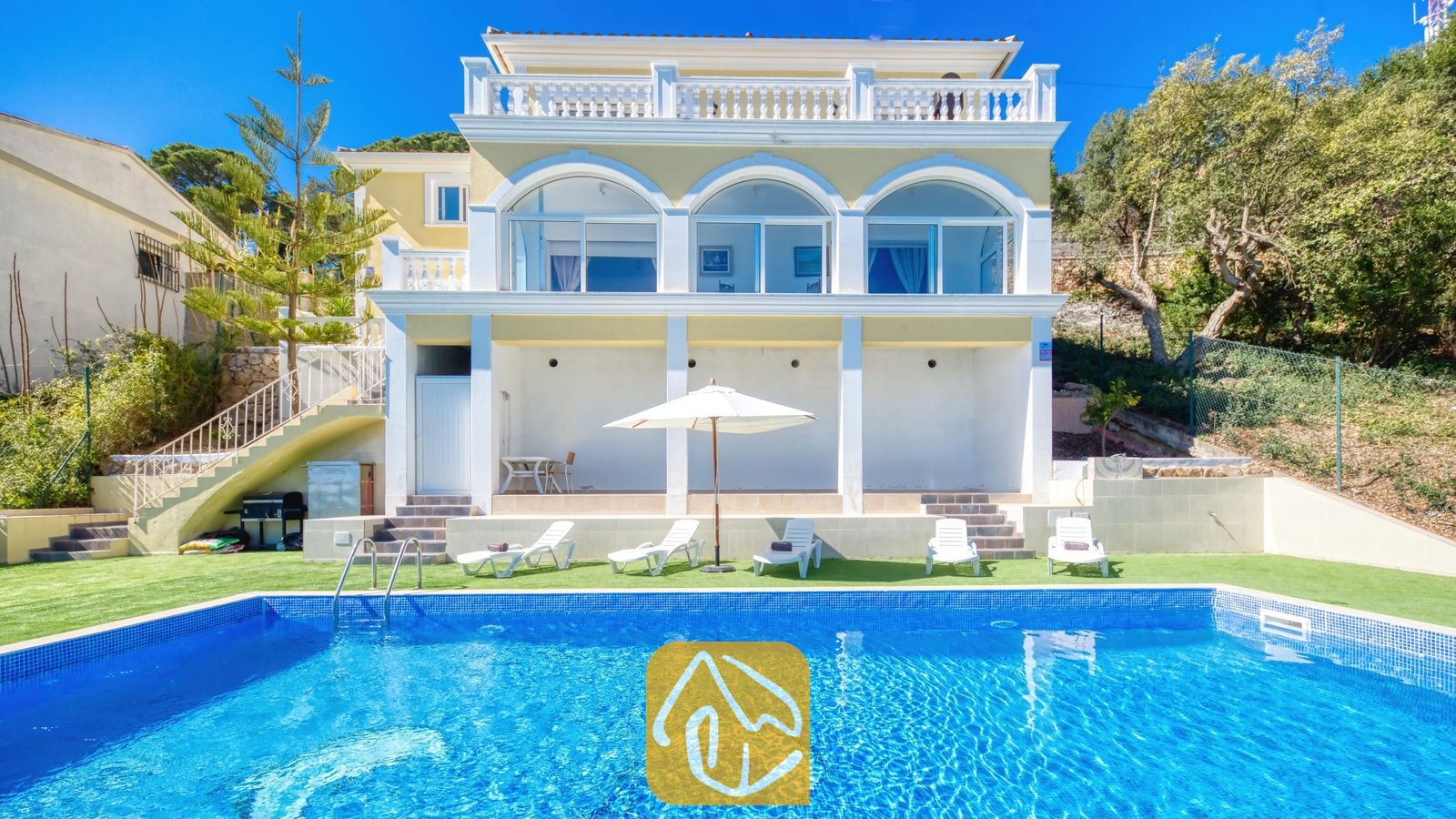 Vakantievilla's Costa Brava Spanje - Villa Sunrise - Buitenkant van Villa Sunrise met een rechthoekig, blauw getegeld zwembad, witte ligbedden en een gele villa van meerdere verdiepingen met gebogen glazen deuren onder een heldere hemel.