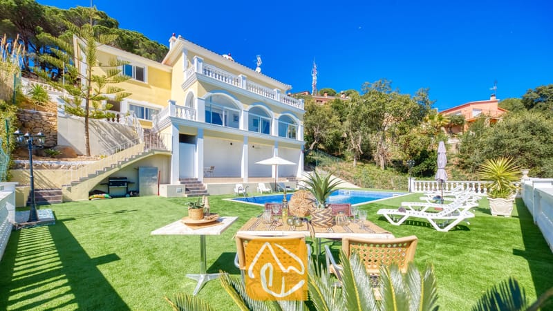 Vakantievilla's Costa Brava Spanje - Villa Sunrise - Buitenaanzicht van Villa Sunrise met een gele gevel en een wit balkon, een eettafel buiten op het groene gazon en een zwembad.