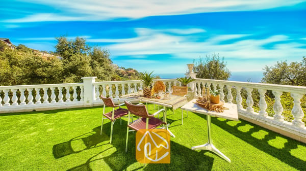 Vakantievilla's Costa Brava Spanje - Villa Sunrise - Terras met een eettafel, geweven stoelen en een witte balustrade met uitzicht op bomen en de blauwe zee bij Villa Sunrise in Spanje.
