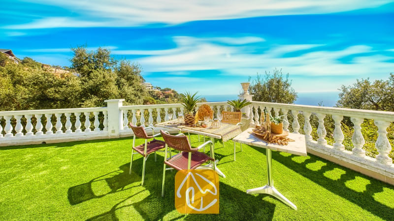 Vakantievilla's Costa Brava Spanje - Villa Sunrise - Terras met een eettafel, geweven stoelen en een witte balustrade met uitzicht op bomen en de blauwe zee bij Villa Sunrise in Spanje.