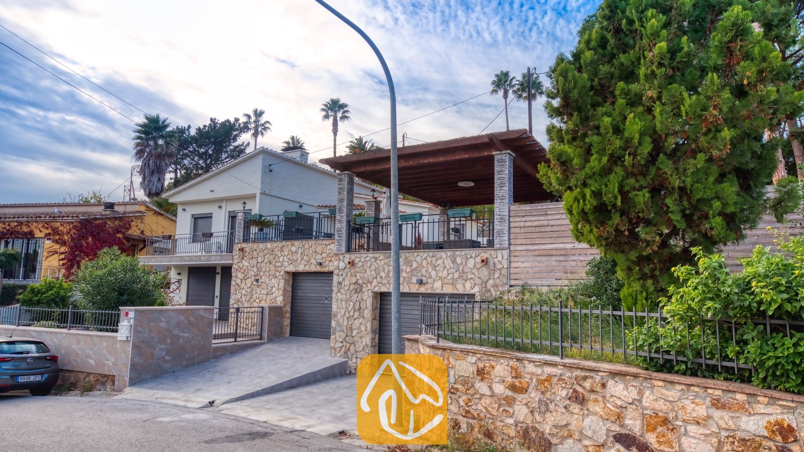 Vakantievilla's Costa Brava Spanje - Villa Palmera - Buitenaanzicht van Villa Palmera met de stenen gevel, de dubbele garagedeuren, de betonnen oprit en het overdekte terras met houten pergola onder een bewolkte hemel.