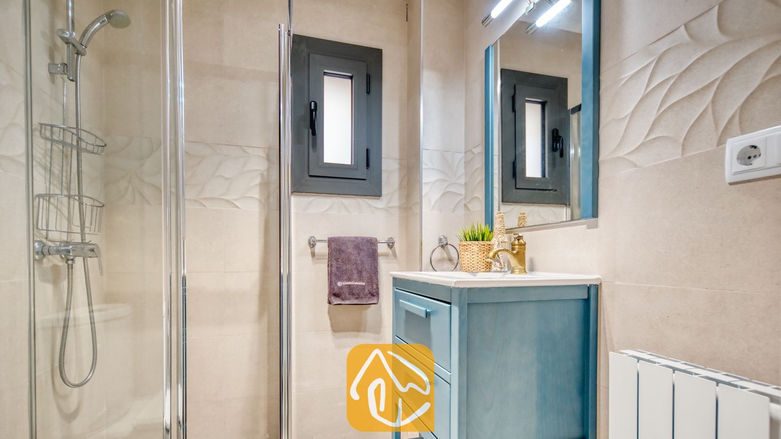 Vakantievilla's Costa Brava Spanje - Villa Palmera - Moderne badkamer met een blauw badkamermeubel en een goudkleurige kraan, een glazen douchecabine en wanden met neutrale, getextureerde tegels.