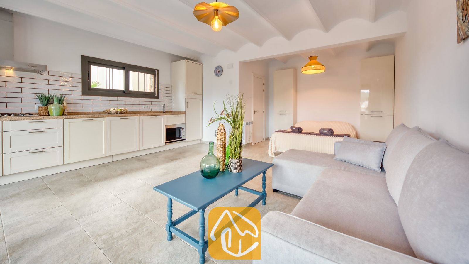 Vakantievilla's Costa Brava Spanje - Villa Palmera - Open woonkamer met een blauwe salontafel, een lichte grijze hoekbank en witte keukenkasten met een tegelachterwand.