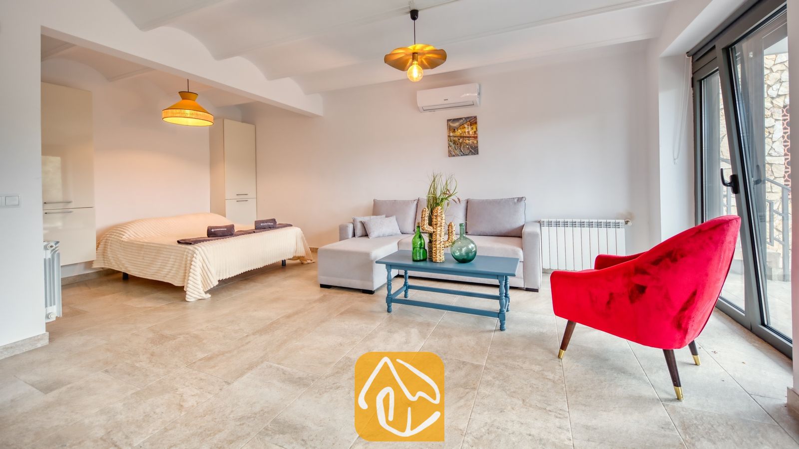 Vakantievilla's Costa Brava Spanje - Villa Palmera - Lichte woonkamer met een grijze hoekbank, een blauwe salontafel, een opvallende rode fauteuil en een aangrenzend bed in Villa Palmera.