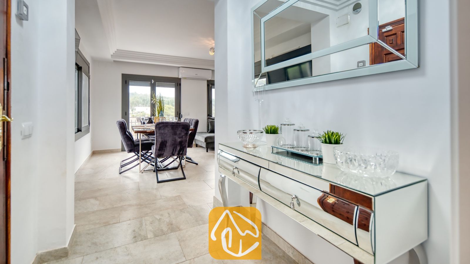 Vakantievilla's Costa Brava Spanje - Villa Palmera - Een consolebord met spiegelafwerking, glazen schalen en kleine potplanten in een hal die leidt naar een eetgedeelte met donkere, gestoffeerde stoelen.