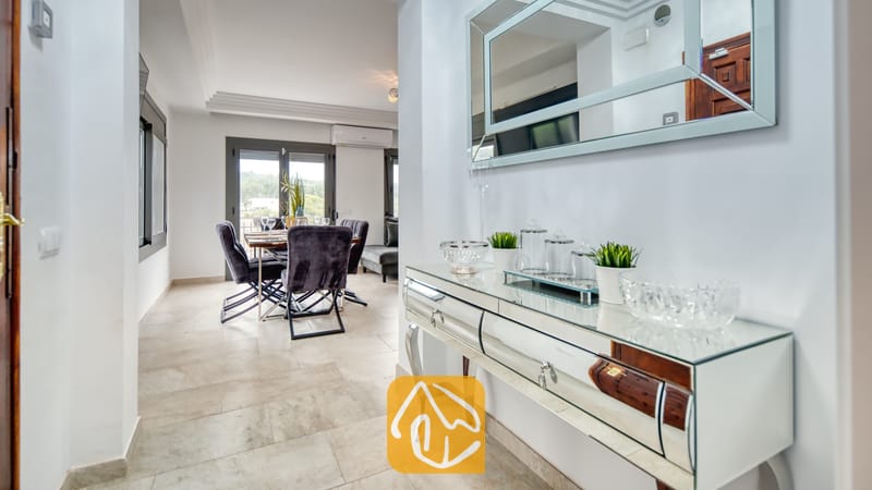 Ferienhäuser Costa Brava Spanien - Villa Palmera - Ein spiegelnder Konsolentisch mit Glasschalen und kleinen Topfpflanzen in einem Flur, der zu einem Essbereich mit dunklen, gepolsterten Stühlen führt.