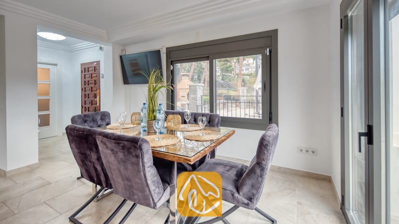Ferienhäuser Costa Brava Spanien - Villa Palmera - Essbereich mit einem glasgedeckten Tisch, umgeben von dunkelgrauen Samtstühlen, platziert vor einem Fenster mit Blick nach draußen.