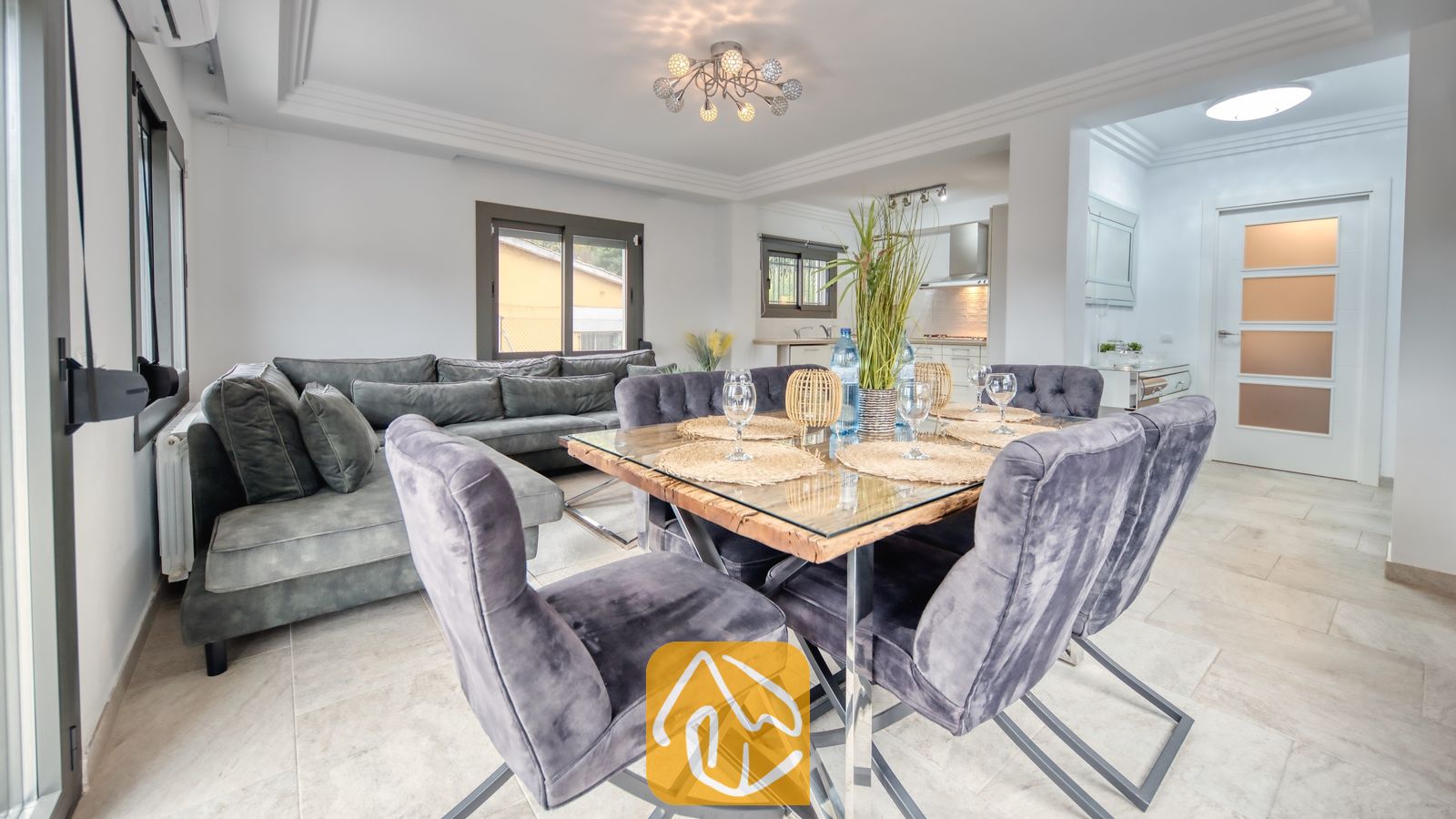 Vakantievilla's Costa Brava Spanje - Villa Palmera - Eethoek voorzien van een houten tafel met glazen blad, omringd door grijs velvet stoelen bij Villa Palmera, met een aangrenzende woonkamer en bank.