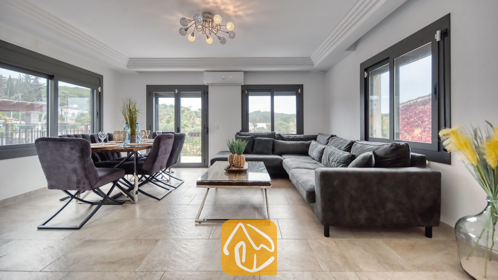 Vakantievilla's Costa Brava Spanje - Villa Palmera - Ruime open woon- en eetkamer in Villa Palmera, voorzien van een grote donkergrijze hoekbank, een houten salontafel en een eettafel met bijpassende fluwelen stoelen.