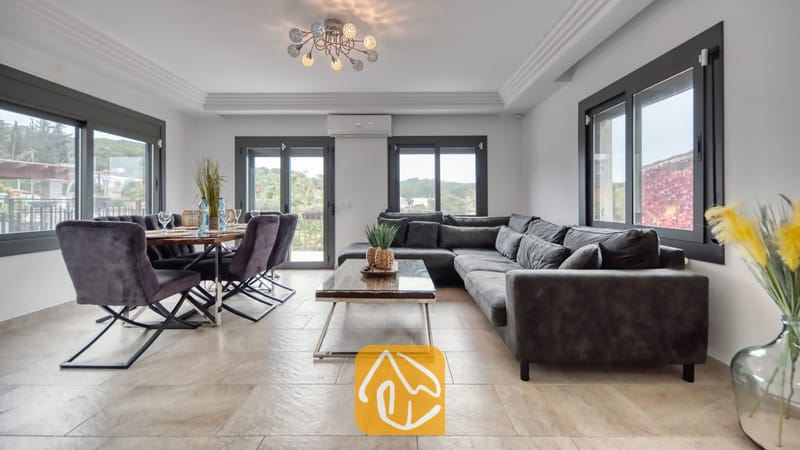 Vakantievilla's Costa Brava Spanje - Villa Palmera - Ruime open woon- en eetkamer in Villa Palmera, voorzien van een grote donkergrijze hoekbank, een houten salontafel en een eettafel met bijpassende fluwelen stoelen.