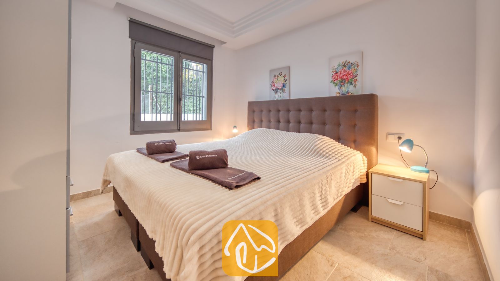 Vakantievilla's Costa Brava Spanje - Villa Palmera - Slaapkamer met een groot bed voorzien van een getufte bruine hoofdbord, wit gestructureerd beddengoed, opgevouwen bruine handdoeken, een nachtkastje met een blauwe lamp en een raam met uitzicht op groen loof