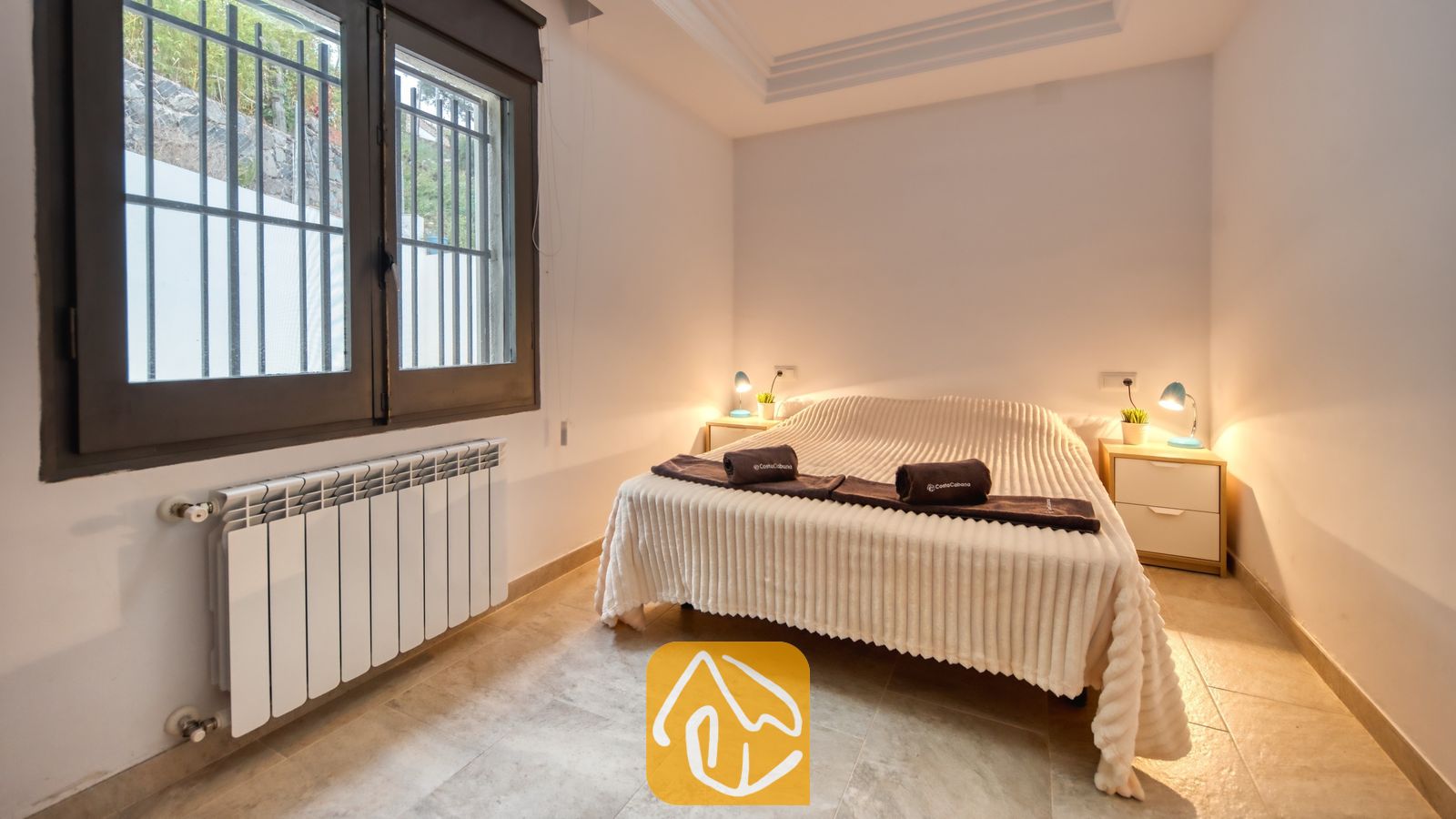 Vakantievilla's Costa Brava Spanje - Villa Palmera - Slaapkamer met een tweepersoonsbed voorzien van een wit sprebed met textuur, opgerolde donkere handdoeken, twee nachtkastjes met kleine lampjes, tegelvloer en een raam met tralies.