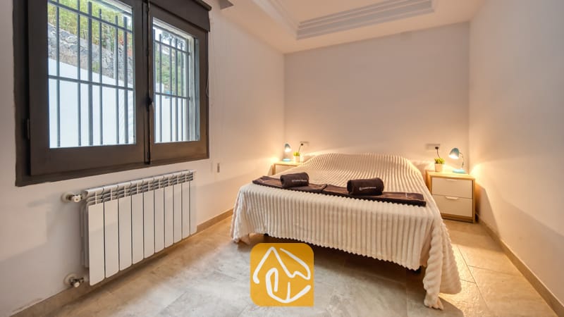 Ferienhäuser Costa Brava Spanien - Villa Palmera - Schlafzimmer mit einem Doppelbett, bezogen mit einer strukturierten weißen Tagesdecke, zusammengerollten dunklen Handtüchern, zwei Nachttischen mit kleinen Lampen, Fliesenboden und einem vergitterten Fenster.
