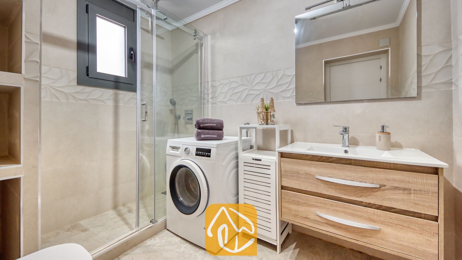Vakantievilla's Costa Brava Spanje - Villa Palmera - Moderne badkamer met een glazen inloopdouche, houten badkamermeubel met een witte wastafel, en een wasmachine met opgevouwen handdoeken erop.